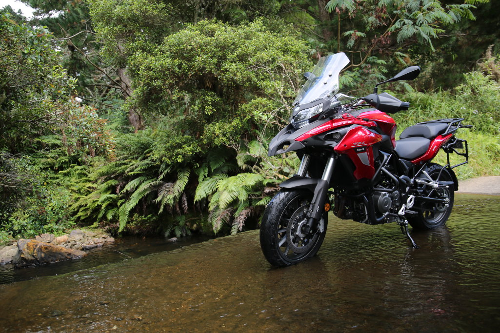عکس موتورسیکلت بنلی قرمزرنگ که در یک محیط جنگلی قرار دارد، در کنارش جنگل و زیر چزخ هایش آب قرار دارد