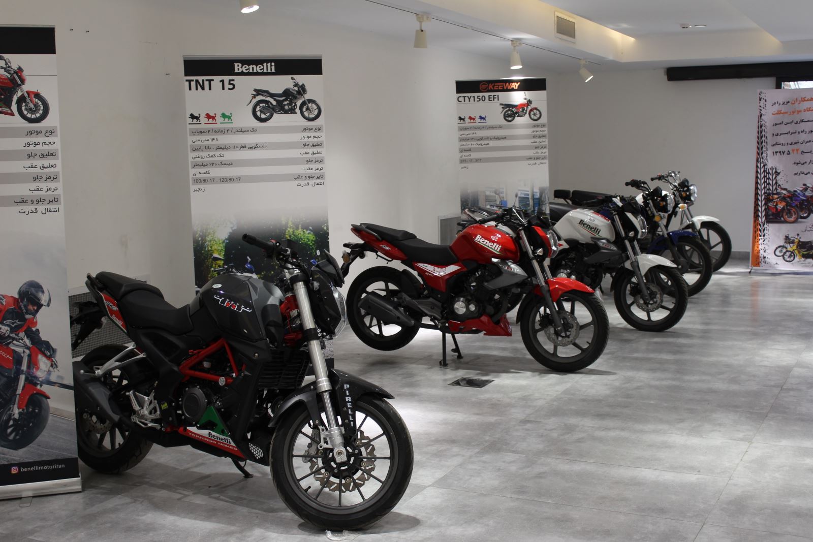 عکسی از سالن نمایشگاه موتورسیکلت که تعداد 5 موتورسیکلت از برند benelli در آن قرار دارد و پشت سر هر موتور یک بنر بزرگ قرار گرفته که عکس، مدل و مشخصات فنی هر کدام در آن نوشته شده.