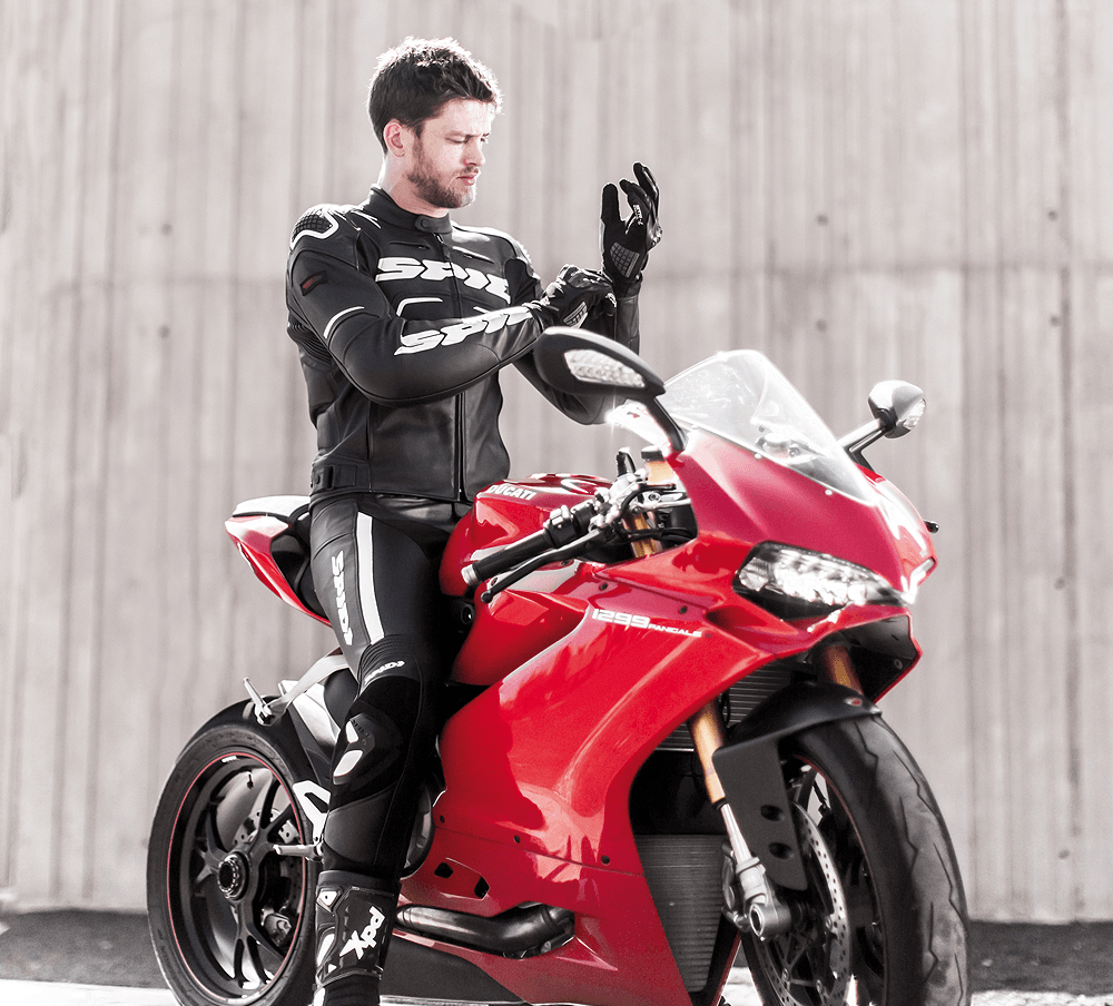 یک مرد جوان با لباس موتورسواری مشکلی که در حال پوشیدن دستکش است سوار بر موتور قرمز رنگ